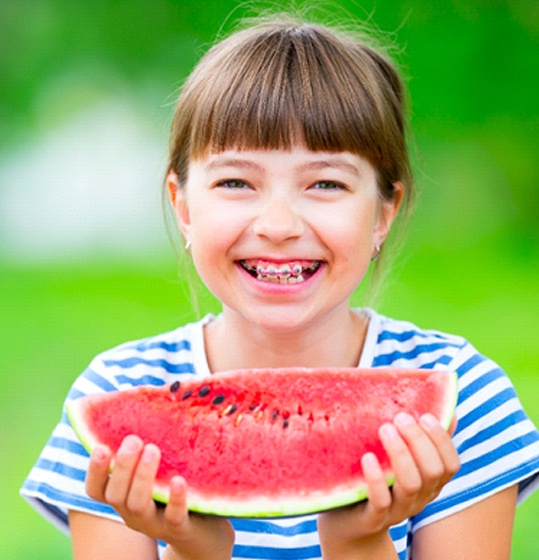 A young girl wearing a striped shirt and smiling with braces preparing to eat a slice of watermelon