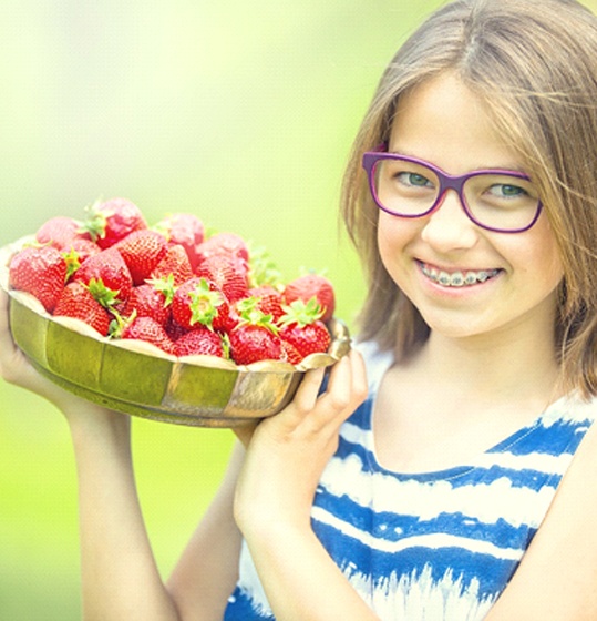 A young girl with purple glasses holding a bowl full of strawberries and smiling while outdoors